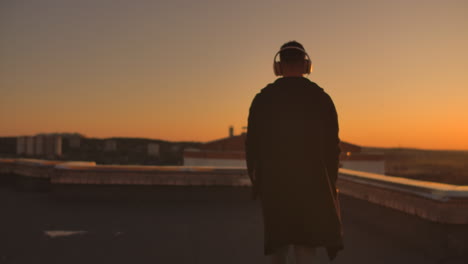 A-man-walks-on-the-roof-at-sunset-with-headphones-looking-at-the-city-from-the-height-of-a-skyscraper-at-sunset.-Relax-while-listening-to-music.-Enjoy-a-beautiful-view-of-the-city-at-sunset-from-the-roof-with-headphones